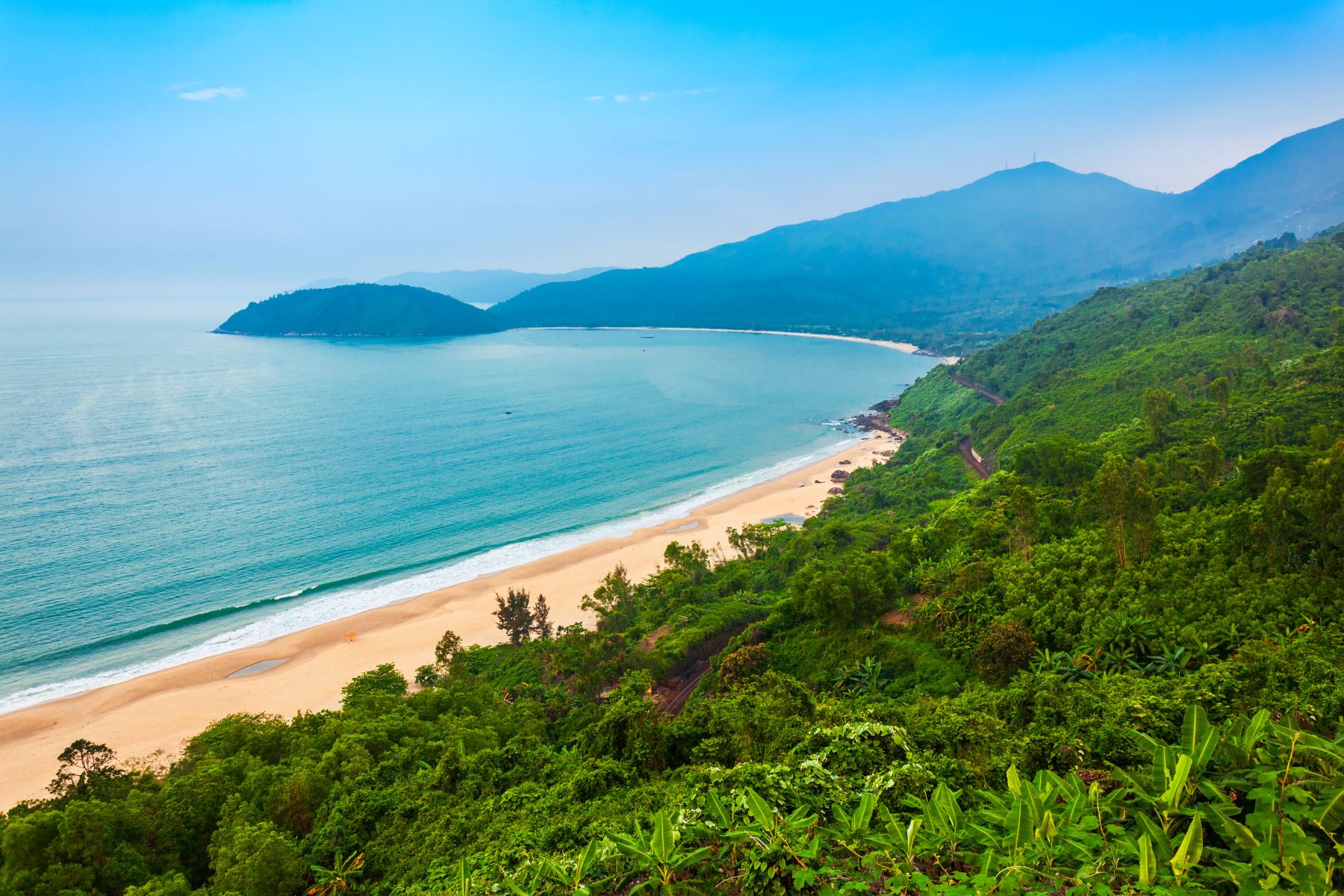 Scenic view of Hai Van Pass featuring lush mountains, dense jungle, a pristine white beach, and an azure bay on a sunny day in central Vietnam