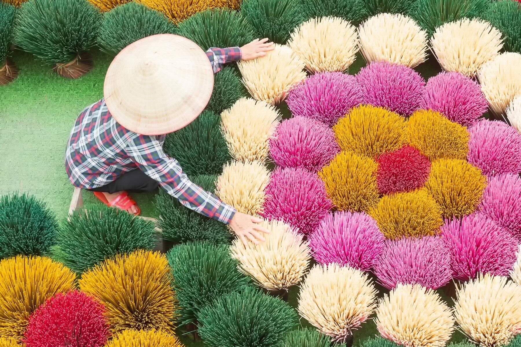 A woman in traditional Vietnamese clothing and a non la hat, surrounded by vibrant incense sticks in pink, white, green, yellow, and red, drying in the sun at Quang Phu Cau Village