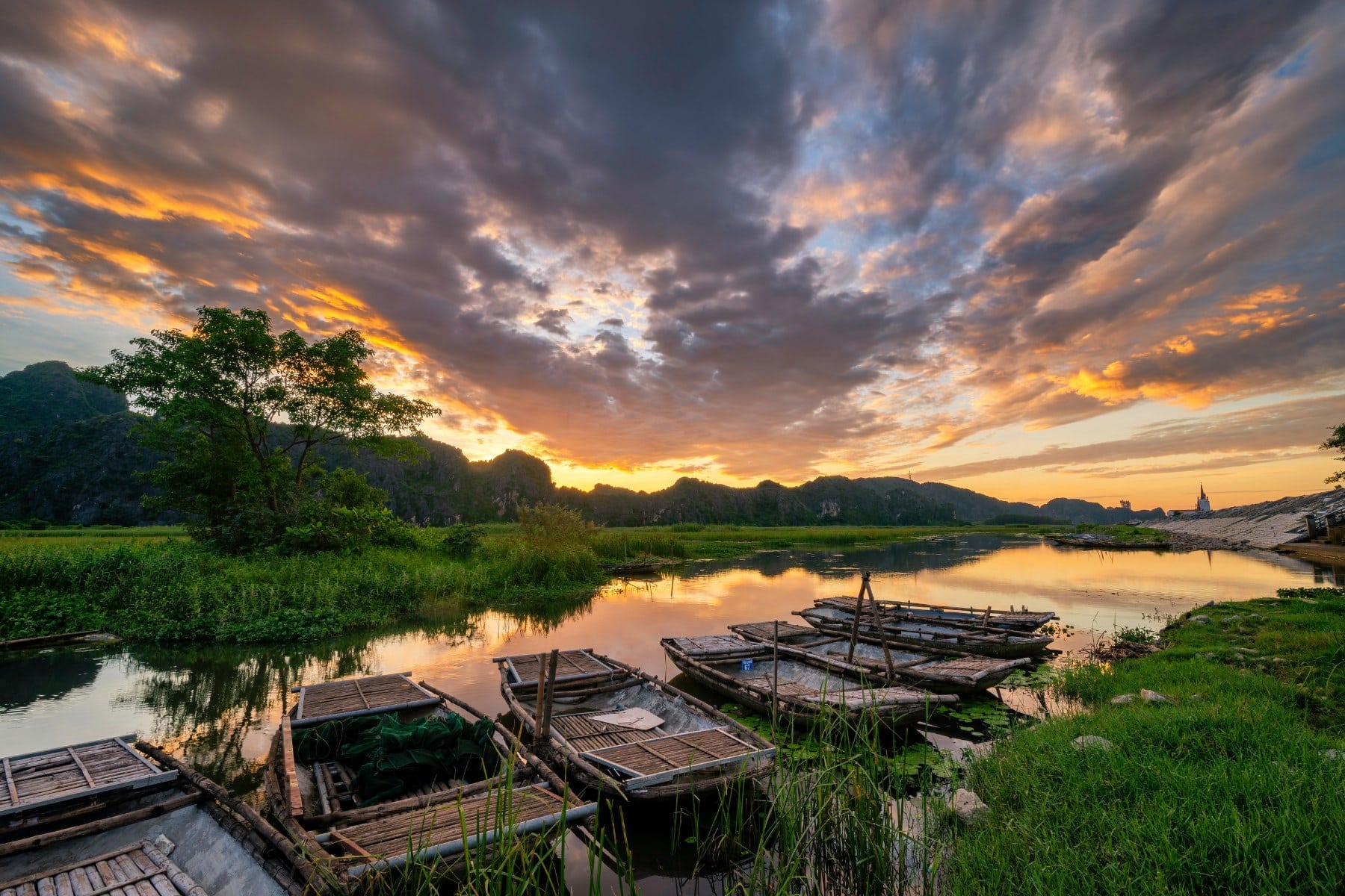 Dawn sunrise over Van Long Wetlands with golden, blue, and grey clouds reflected on the water, and traditional boats docked by the shore