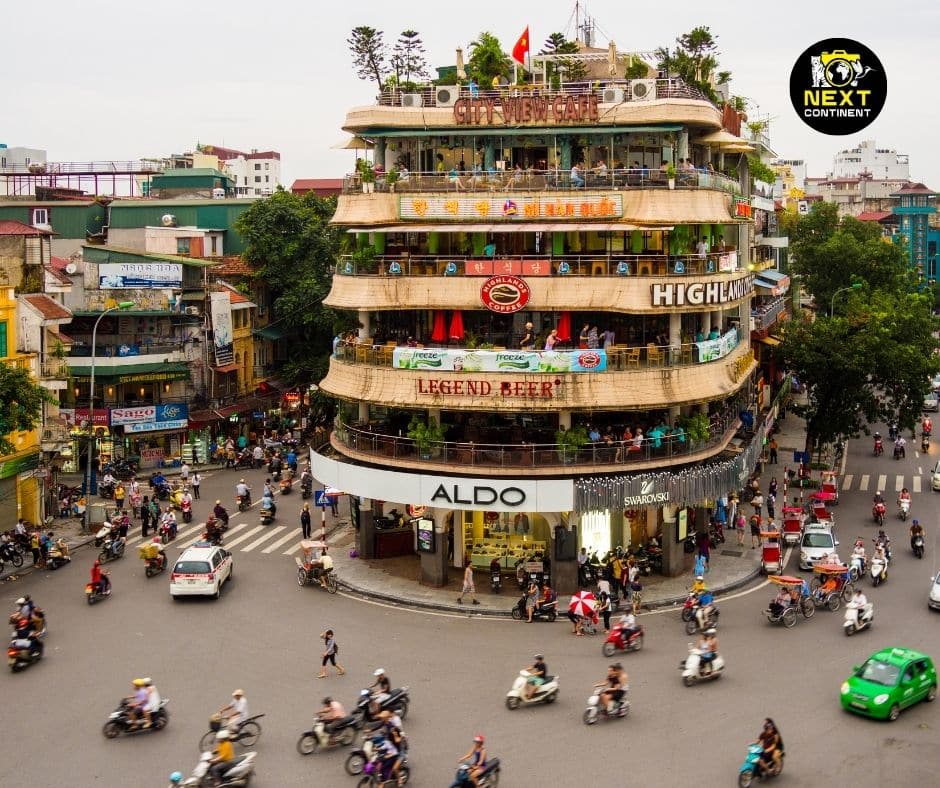 A bustling intersection in Hanoi, Vietnam, with the iconic City View Café building at the center, surrounded by motorbikes and pedestrians navigating the streets.