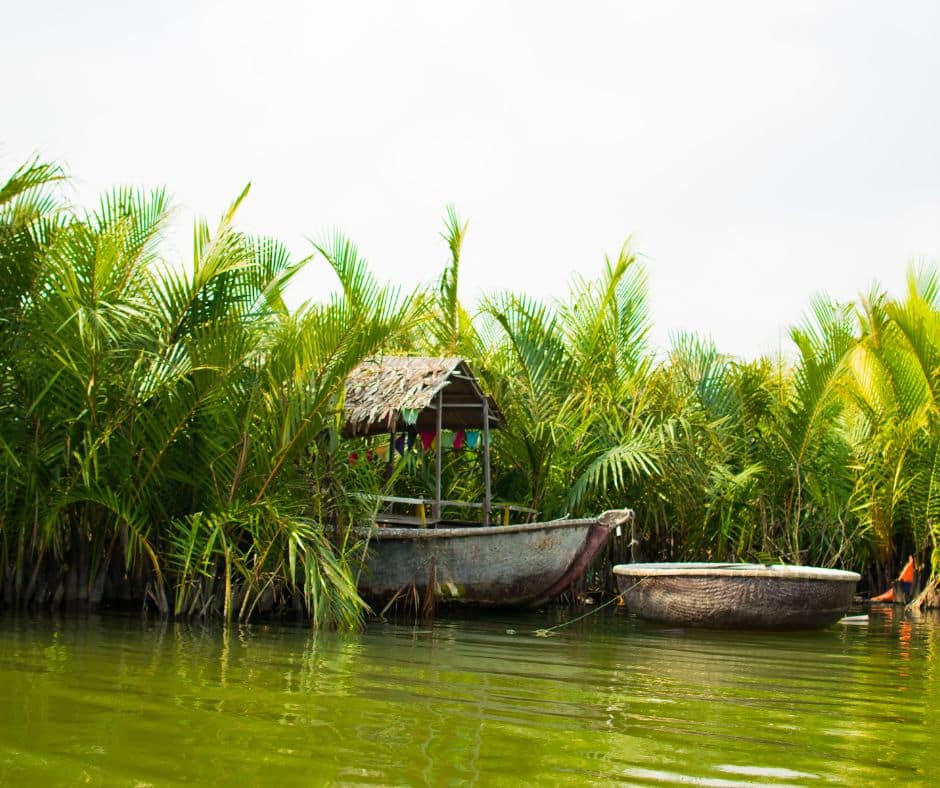 A small wooden boat floating on the calm waterways of Bay Mau Coconut Forest, with a traditional house nestled among lush green palm trees.