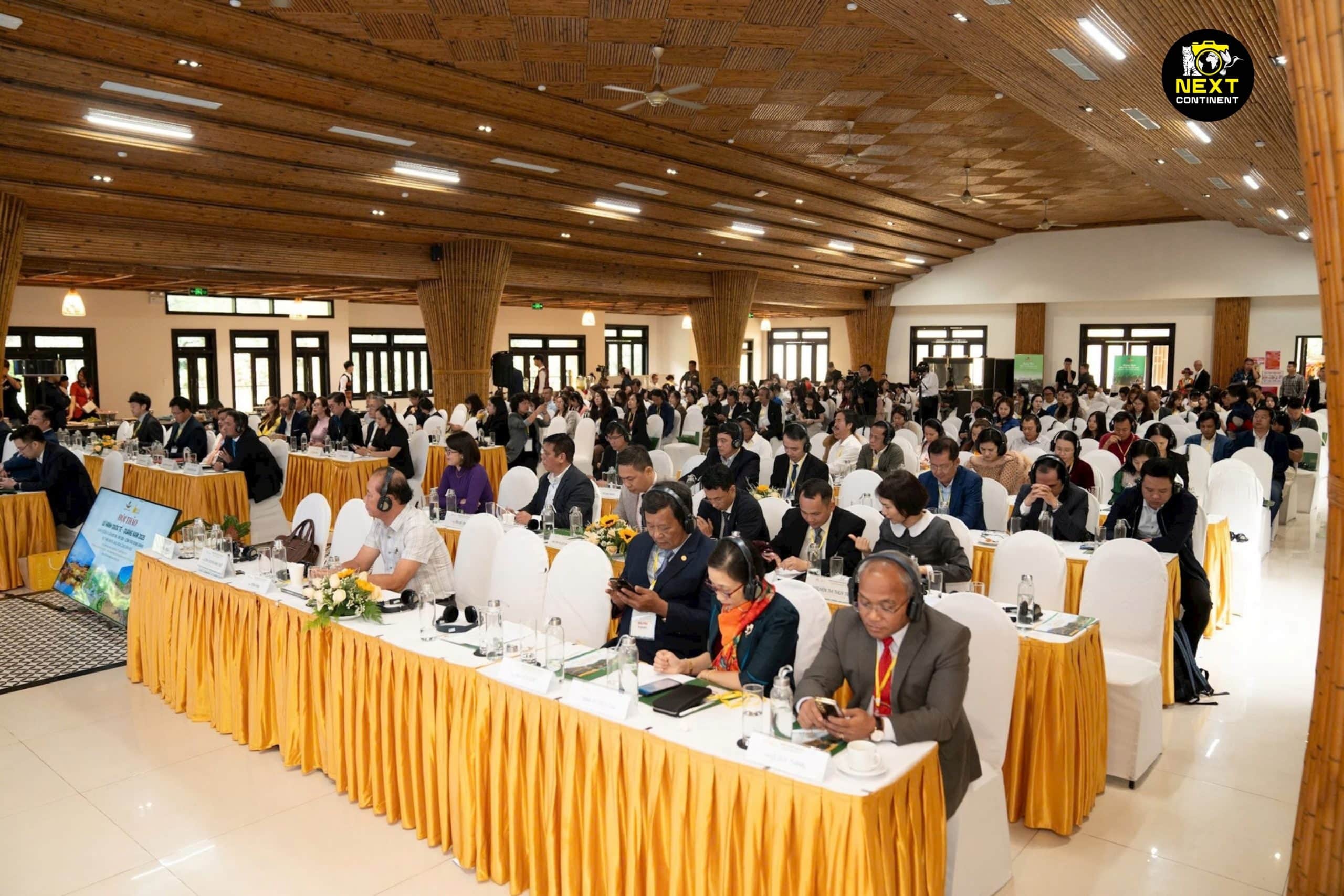 A large international travel conference in Quảng Nam, Vietnam, with over 300 delegates from various countries. Attendees are seated in rows, listening to speakers, taking notes, and using translation headsets. The event is held in a spacious hall with wooden decor, reflecting the region’s cultural heritage.
